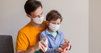 Father helping son use antibacterial gel on his hands. Both wear surgical masks.