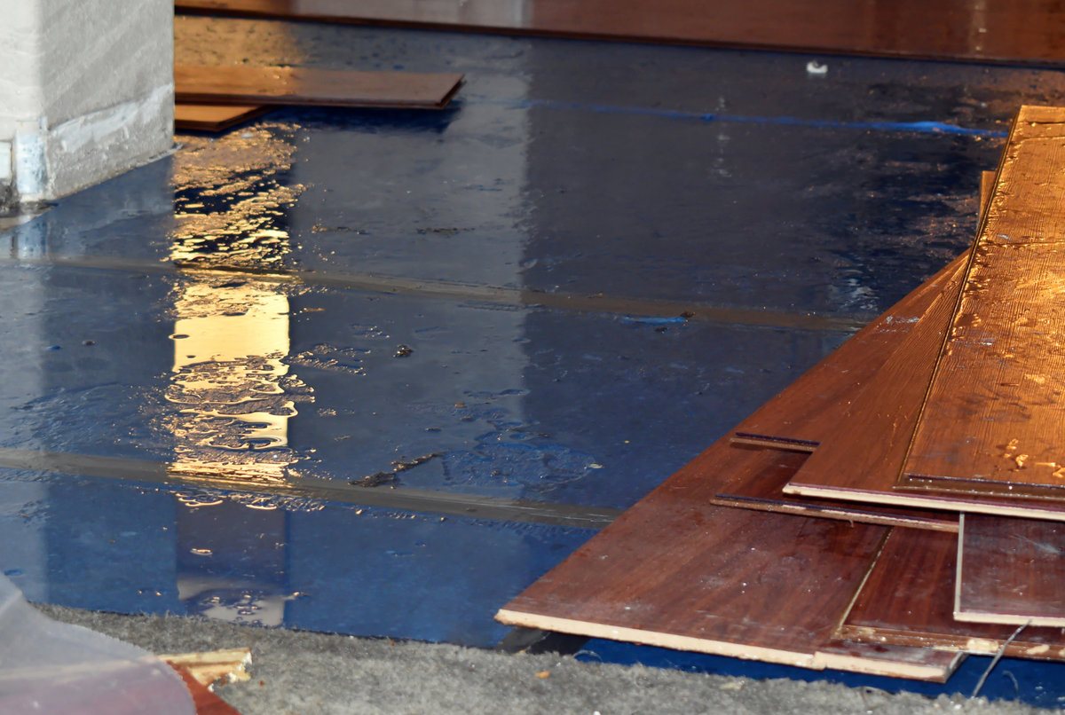 Water soaking concrete floor in an unfinished basement with medium brown wood floor planks piled up on top of each other. 