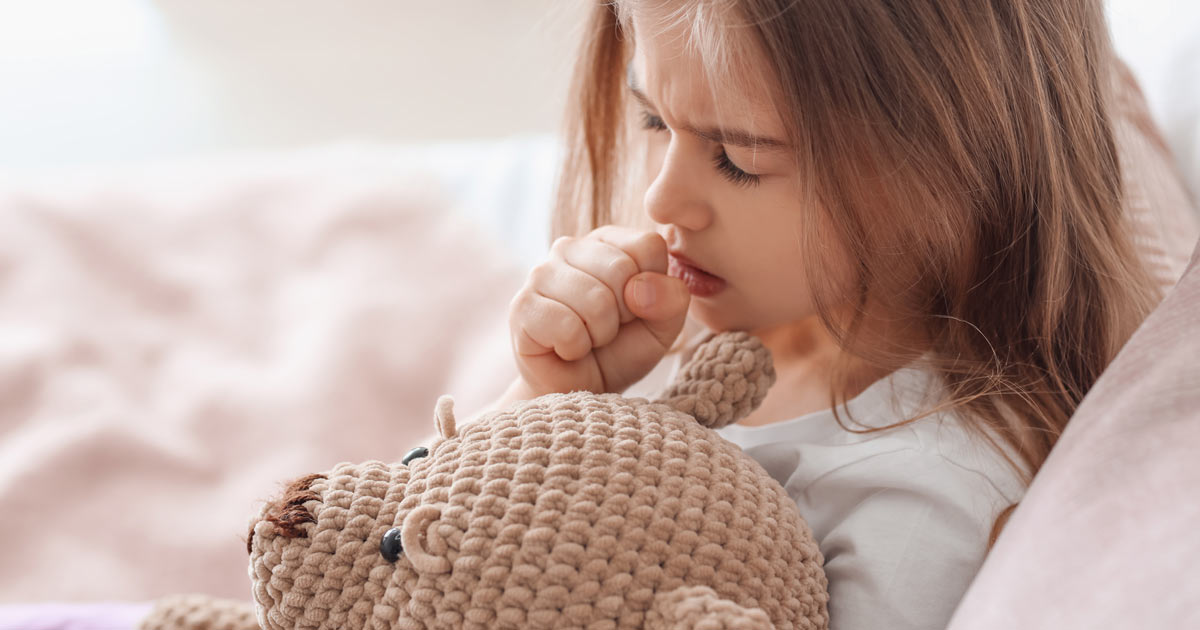Little girl in bed with her teddy bear and coughing into her fist.