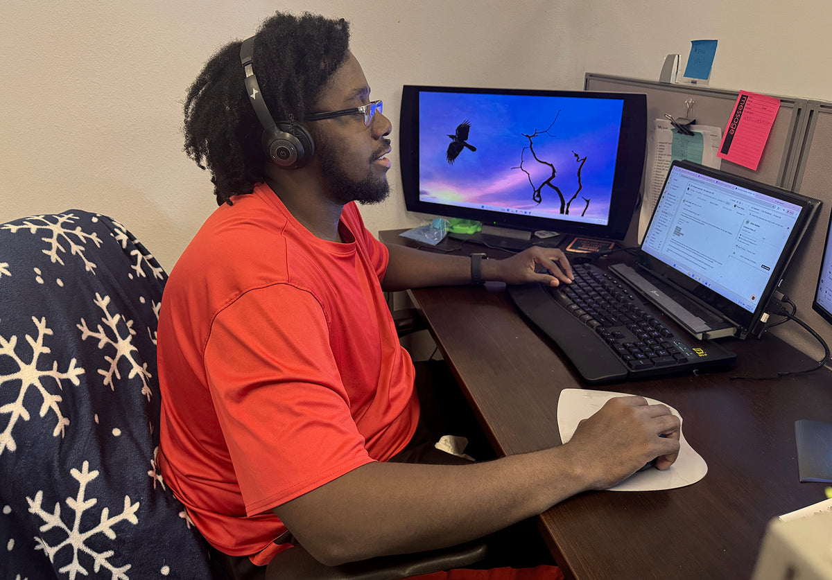 A man with dark curly hair, glasses, and a beard sits at a desk wearing a bright orange t-shirt and headphones, working on a laptop and computer monitor in an office setting.