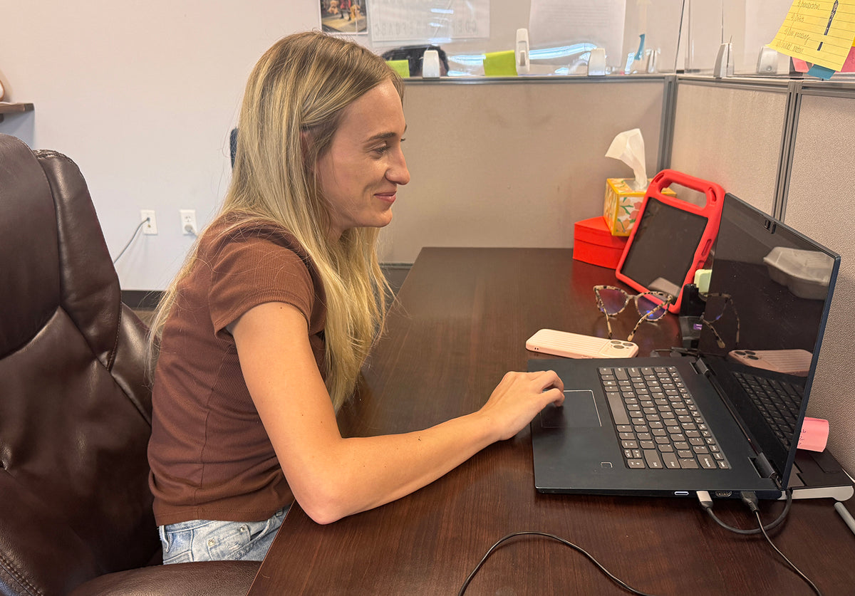 A woman with blonde hair sits at a cubicle desk using a black laptop. She is wearing a brown t-shirt and light blue jeans.