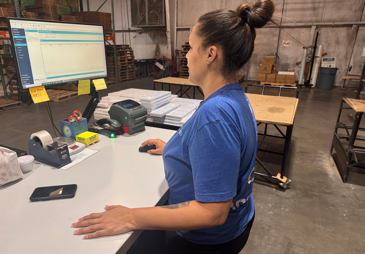 A woman in a blue t-shirt stands at a counter in a warehouse setting, using a computer mouse in front of a computer monitor.