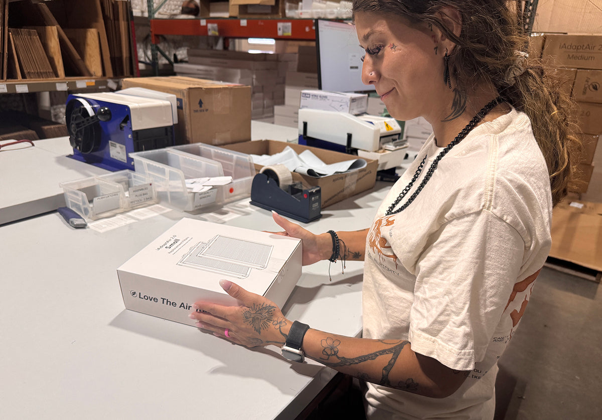A woman in a white graphic t-shirt stands at a table in a warehouse setting, holding a white product box with the text '# Love The Air' printed on the side.