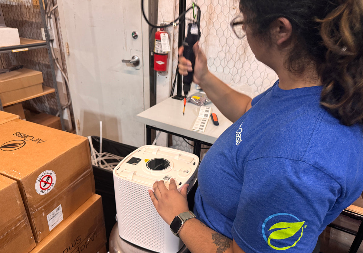 A person in a blue t-shirt uses a handheld device next to a white portable air cleaner in a storage or office environment with cardboard boxes and shelves.