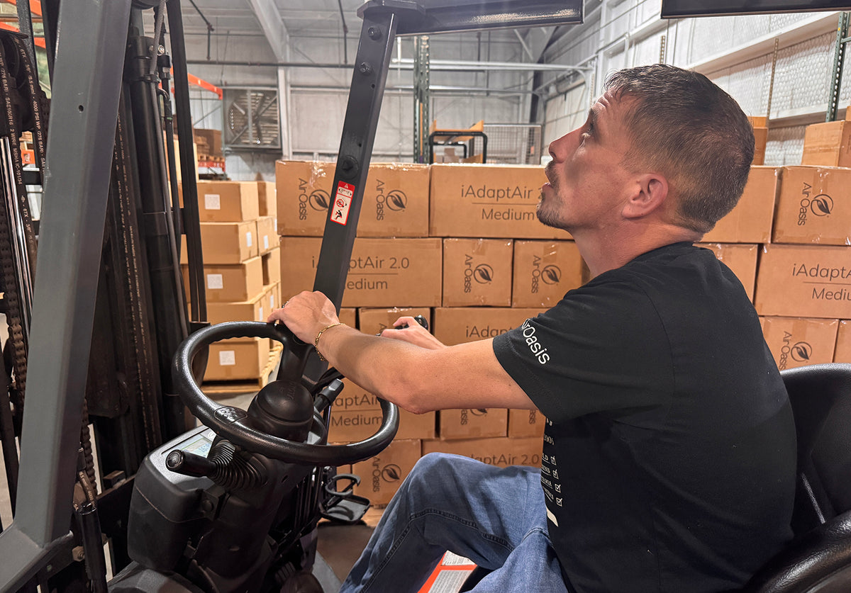 A man wearing a black t-shirt and blue jeans sits in the driver's seat of a forklift, looking up and to the side. Behind him are stacks of cardboard boxes, including some labeled 'iAdaptAir 2.