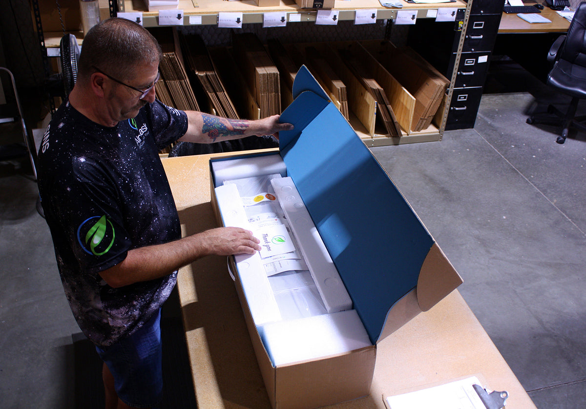 A man in a black galaxy-print t-shirt opens a large cardboard shipping box with white foam padding and blue inner panels in a warehouse setting.