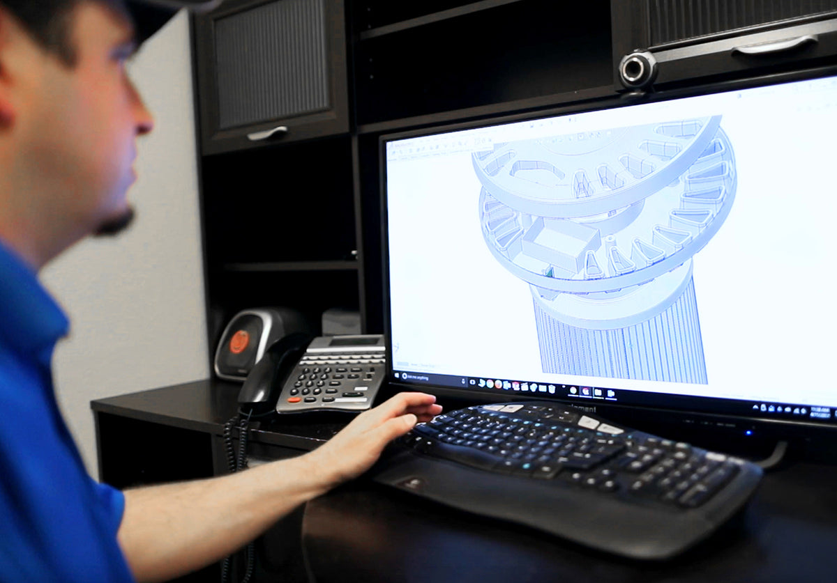 A man in a blue shirt works at a desk, looking at a computer monitor displaying a blue 3D architectural model of a column. A keyboard and phone sit on the desk in front of him.
